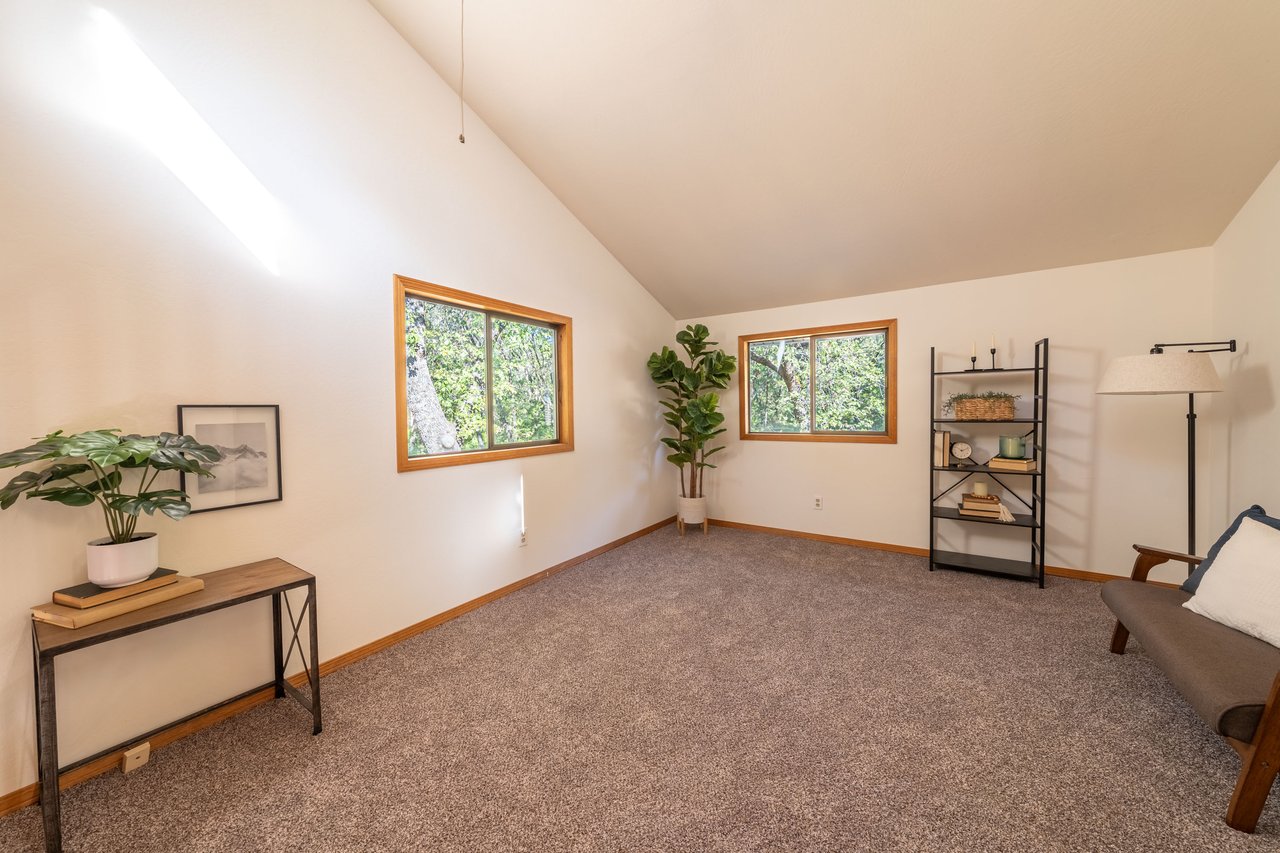 Staged living room with a vaulted ceiling, beige carpet, and windows overlooking leafy trees.