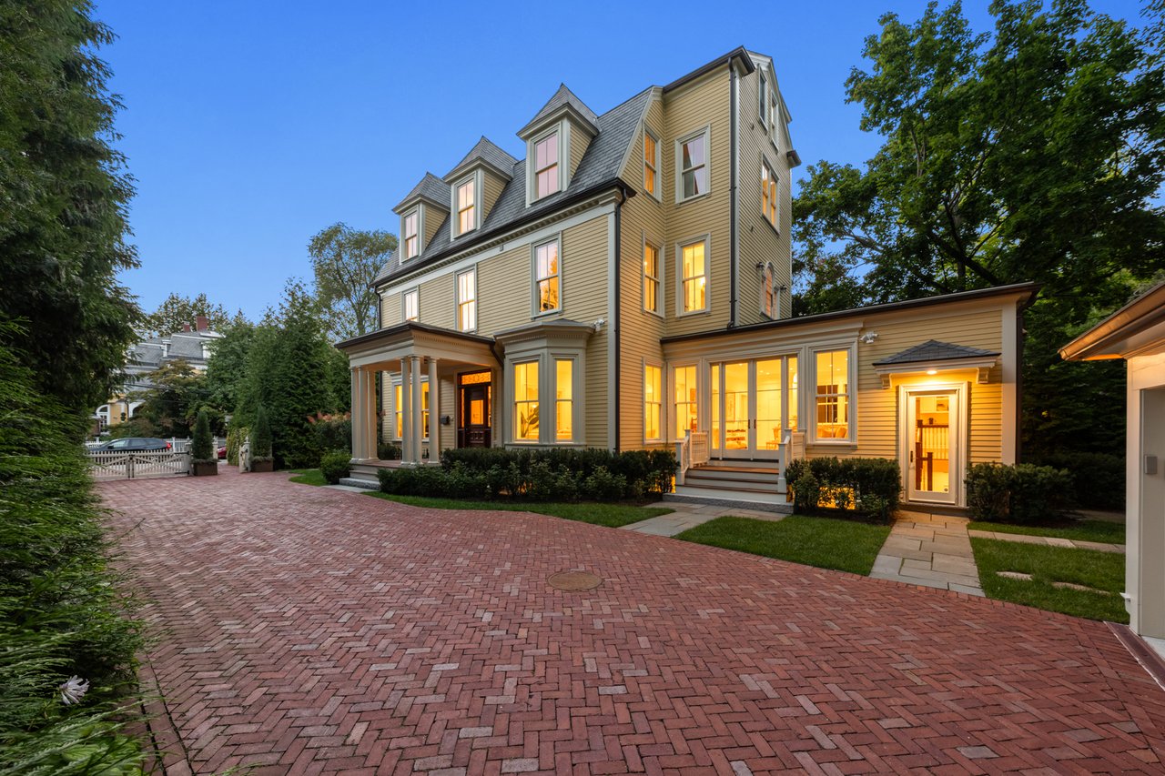Multi-story Victorian house with glowing windows and a broad brick driveway at dusk.