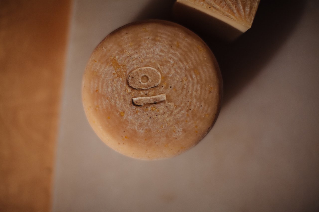 Round artisan cheese with a small stamped mark on its rind, beside another cheese.