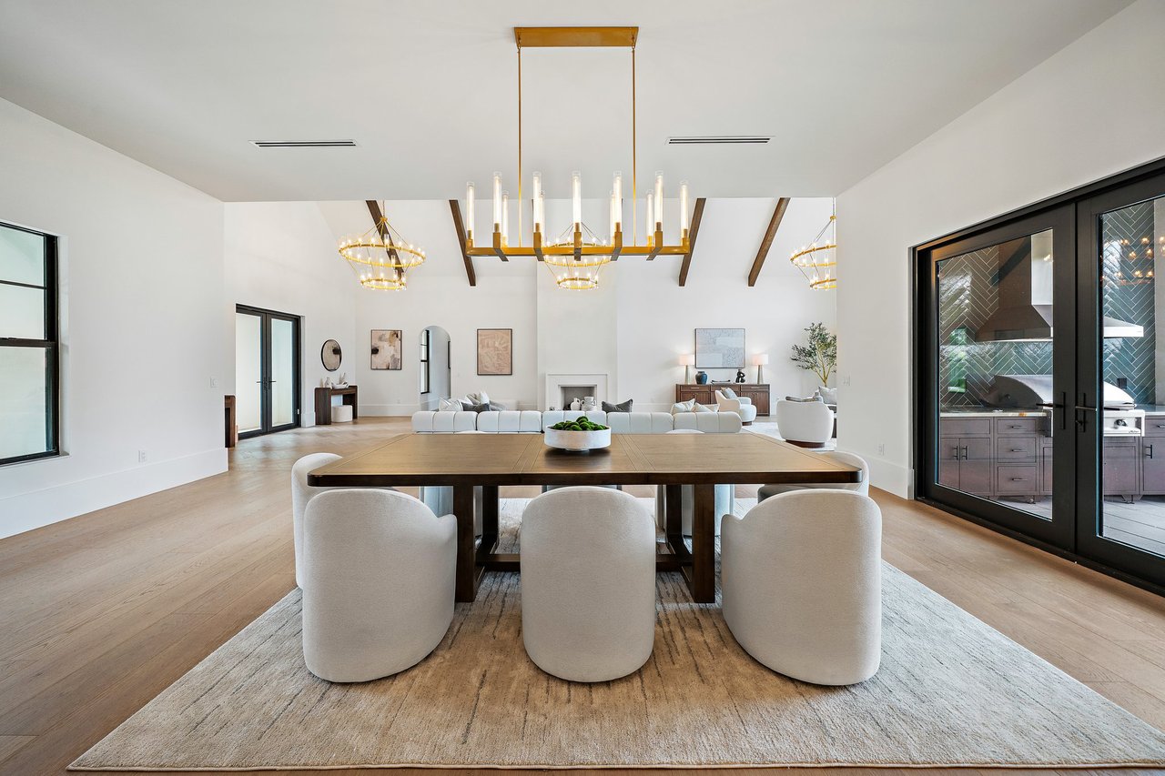 Dining room with a wood table, brass chandelier, exposed beams, and glass doors to an outdoor kitchen.