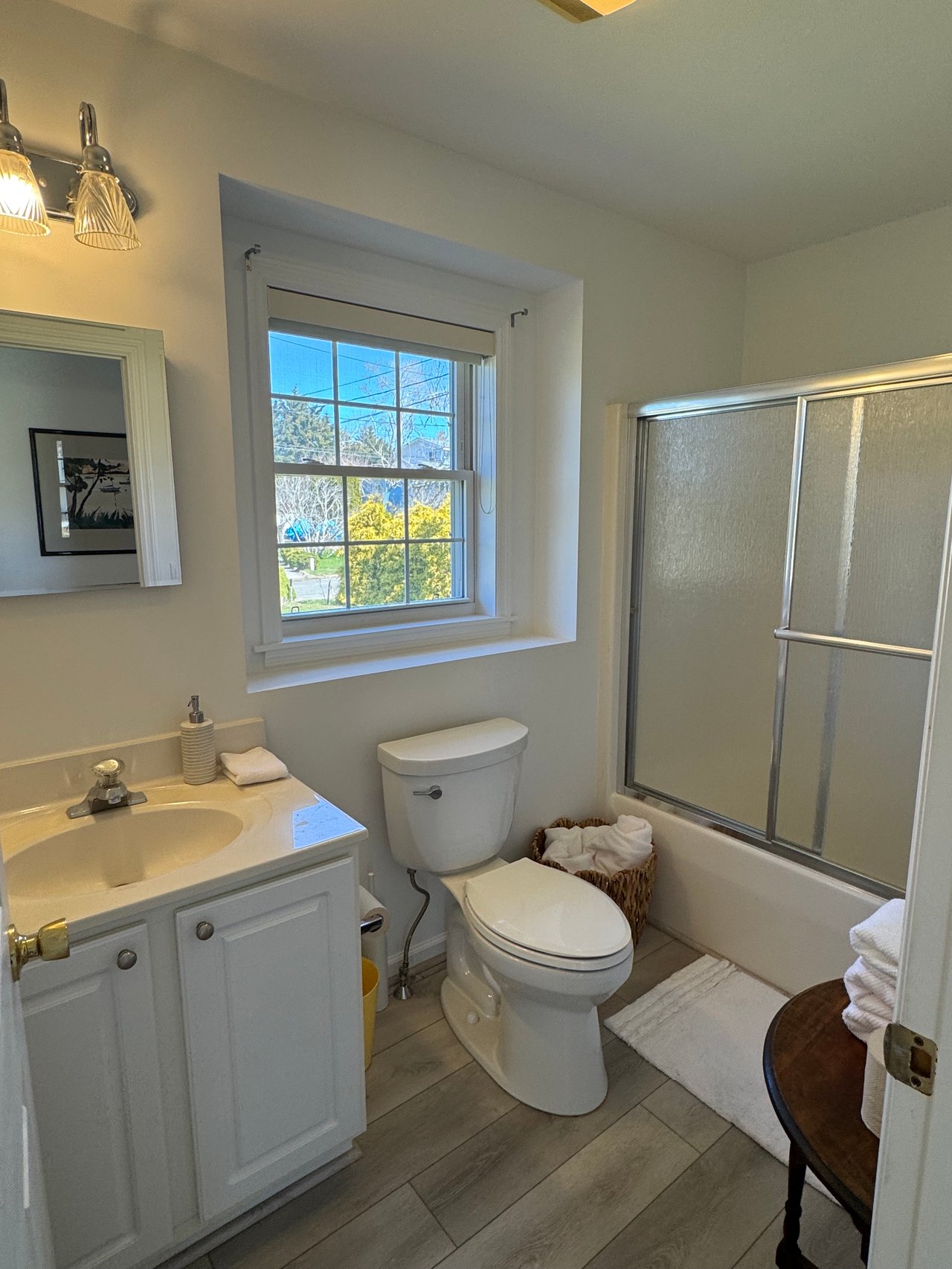 Bathroom with a white vanity, toilet, and glass-enclosed tub beneath a window overlooking trees.