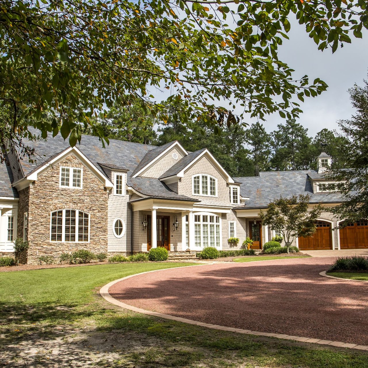 Large stone-and-shingle house with a curved driveway and mature trees overhead.