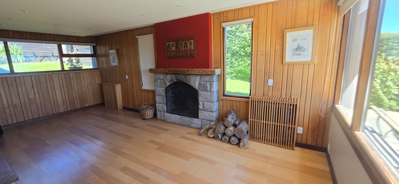 Unfurnished wood-paneled living room with a stone fireplace and large windows overlooking a green yard.