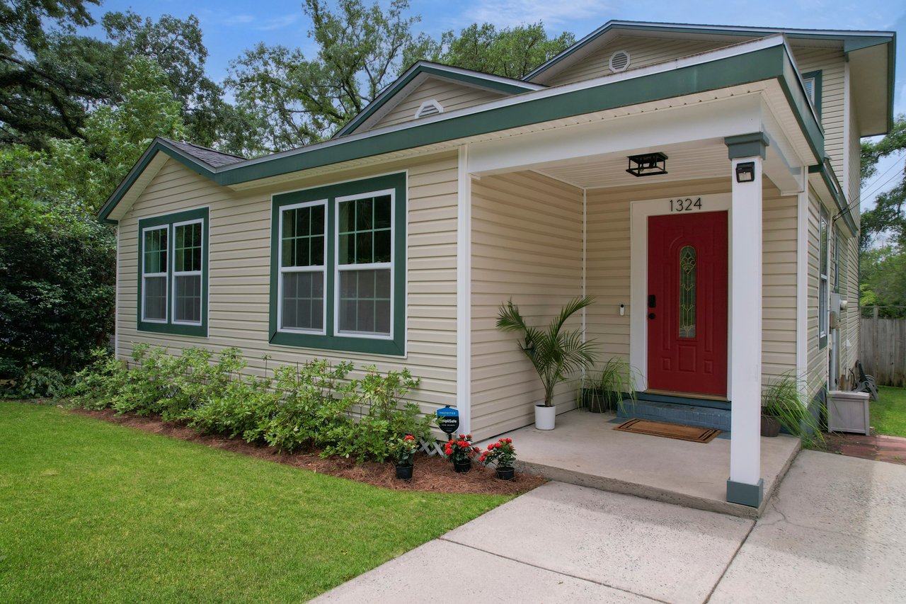 Small cream-colored house with green trim, a red door, and a porch. Surrounded by green grass and shrubs, evoking a welcoming and cozy feel.