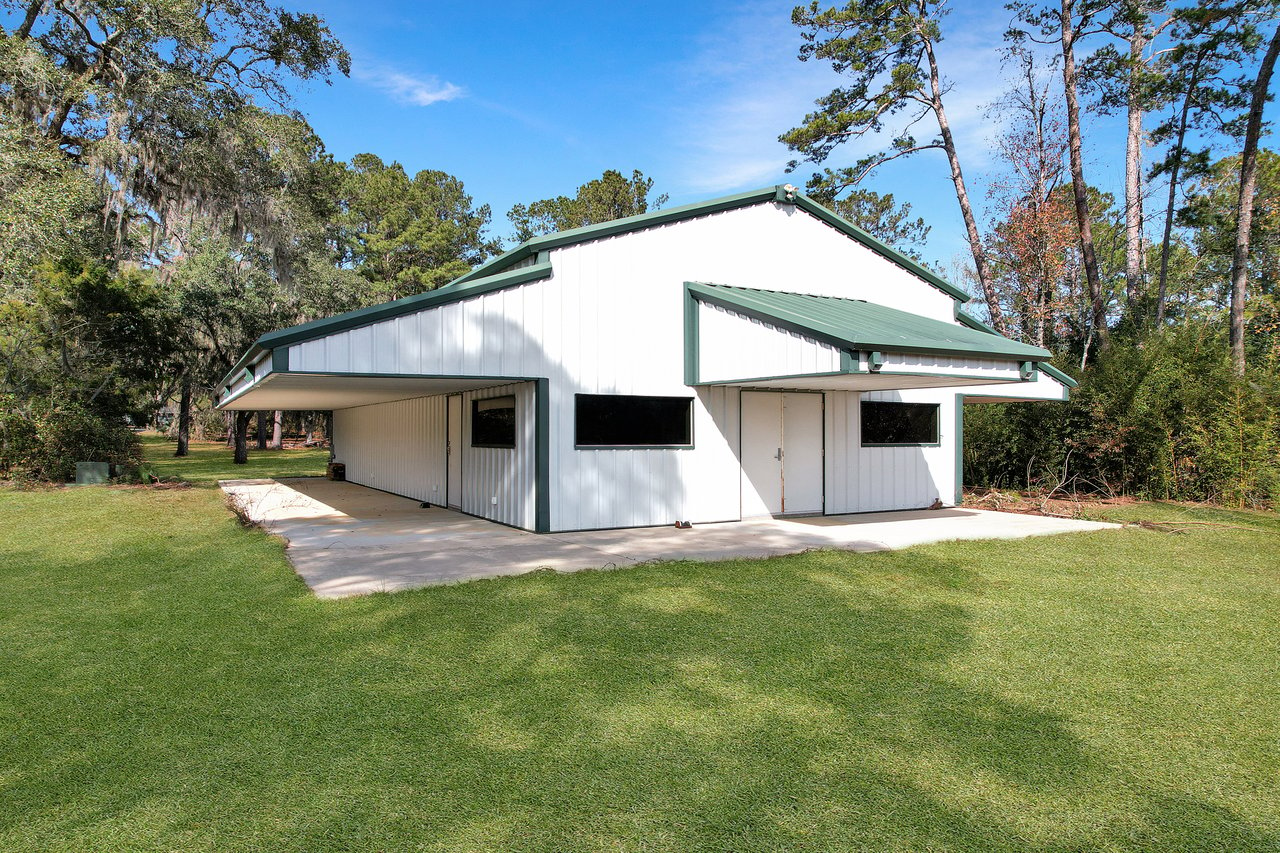 A modern white metal building with a green roof stands on a grassy lawn, surrounded by trees under a clear blue sky, conveying tranquility.