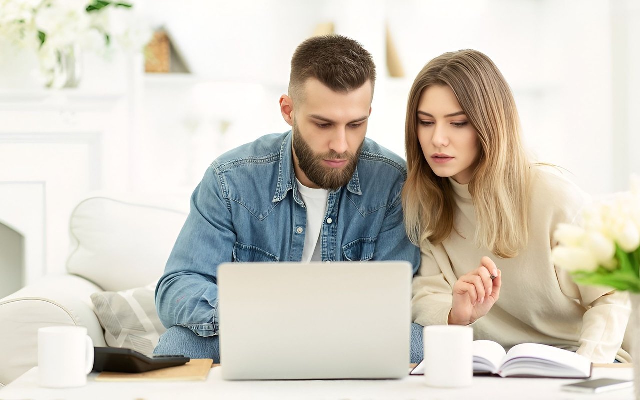 Couple searches on a laptop together on a white sofa, with a tablet and open book nearby.