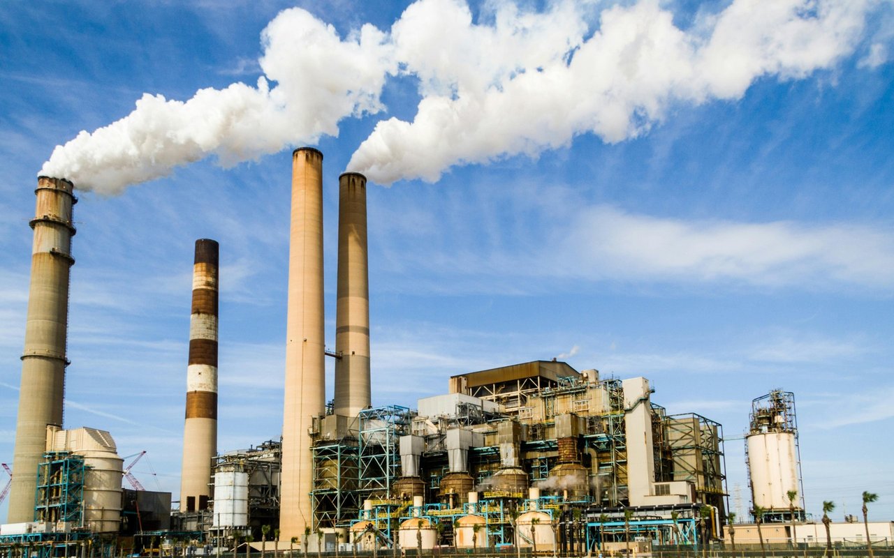 Industrial power plant smokestacks release thick white emissions above processing equipment.