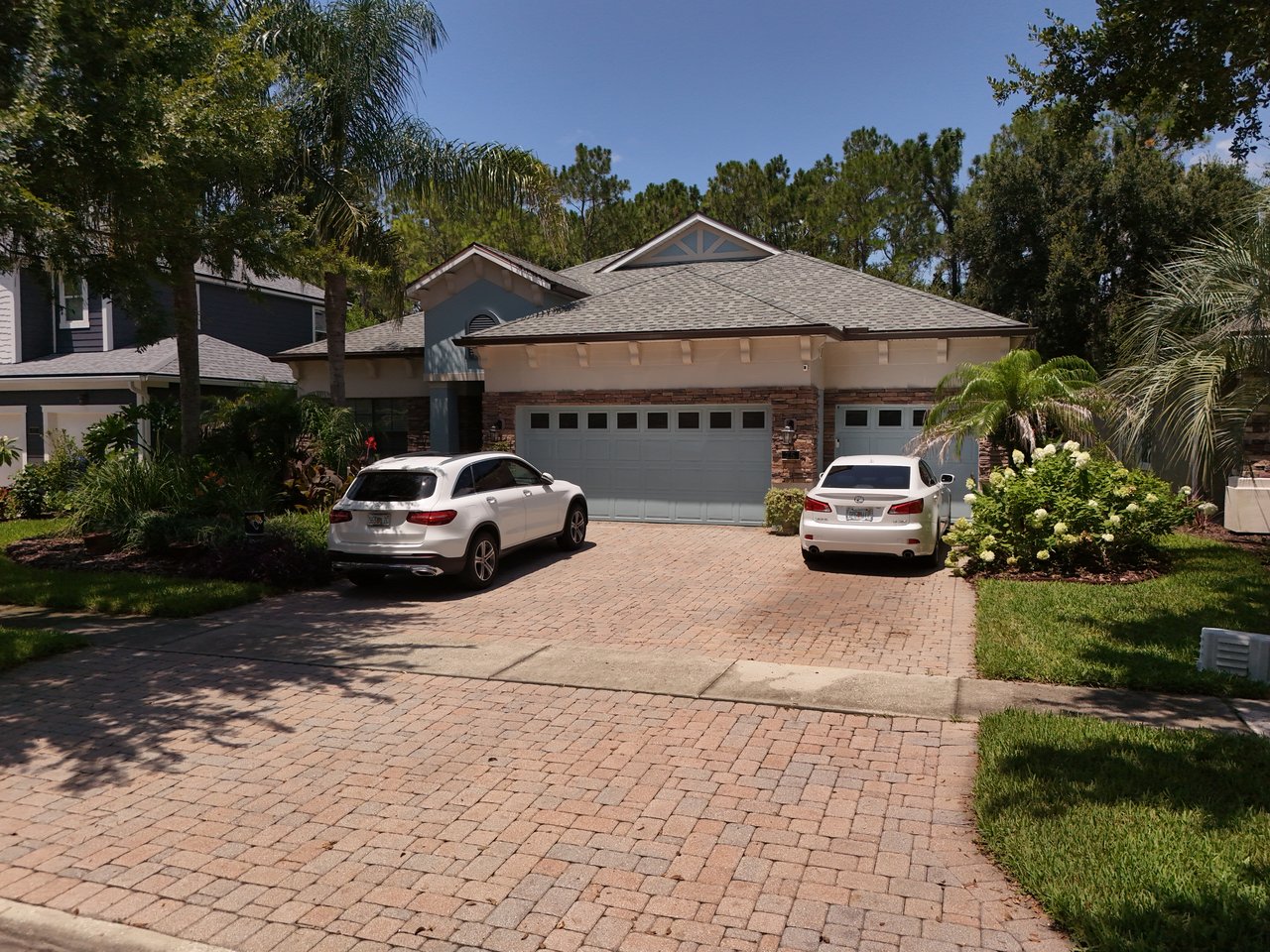 Single-story home with two garage doors, a brick-paver driveway, parked cars, and landscaped palms.