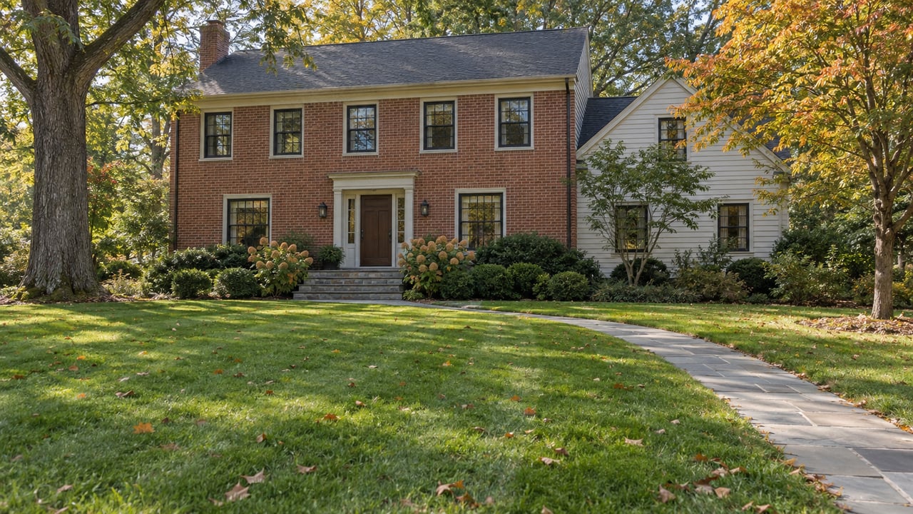 Two-story colonial home with red brick center, clapboard wing, curving stone walk, and broad autumn lawn.