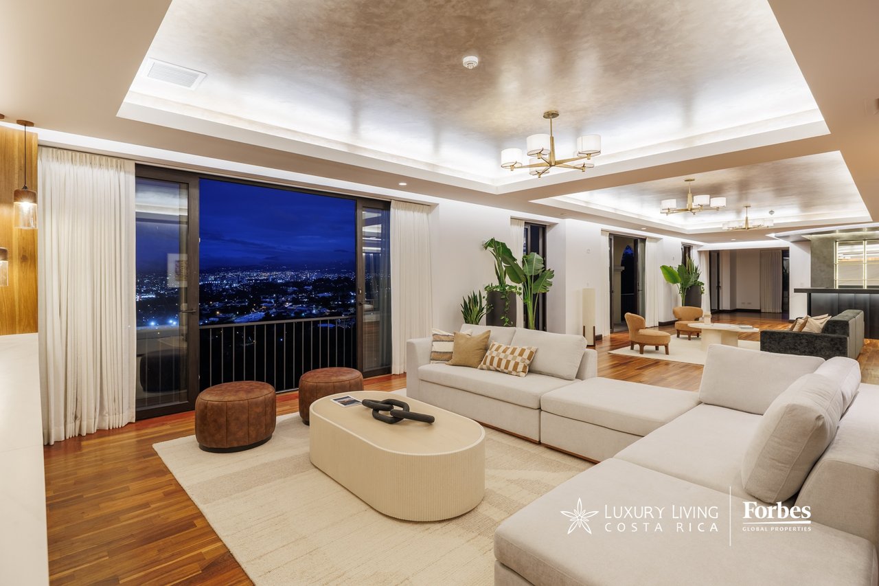 Furnished penthouse living room with white sectional sofas and city lights visible through expansive windows at night.