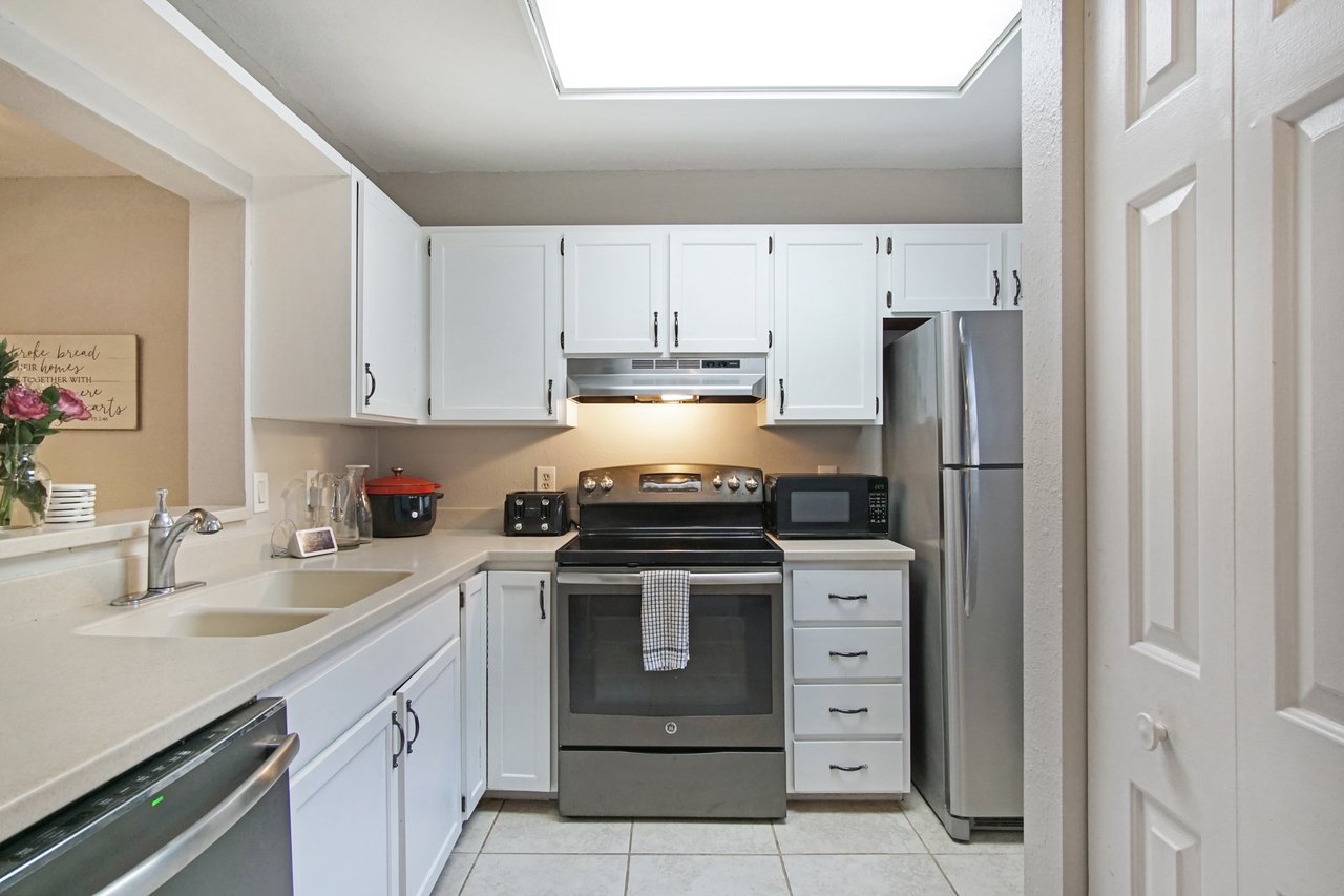 Bright kitchen with white cabinets and stainless steel appliances, including an oven, microwave, and refrigerator. Flowers on the counter add warmth.