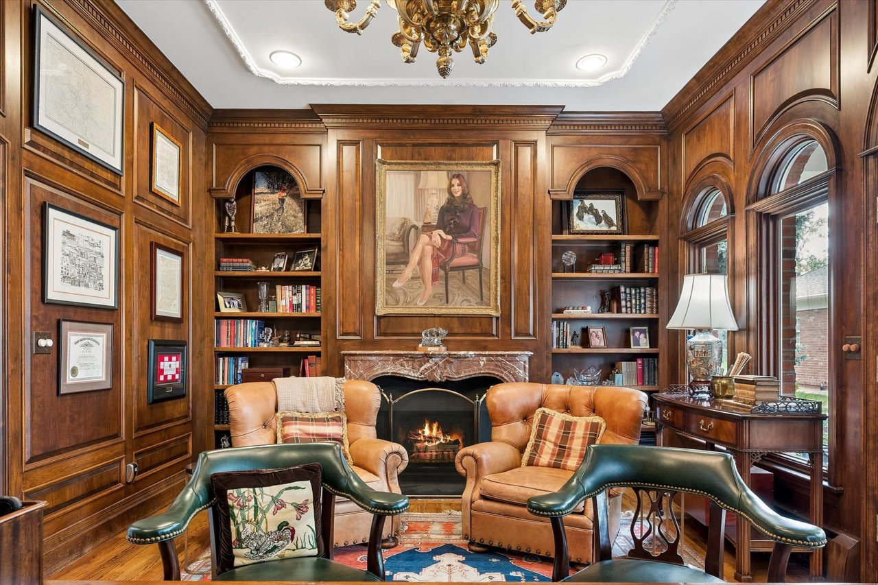 Wood-paneled library with built-in bookshelves, a marble fireplace, leather chairs, and arched windows.
