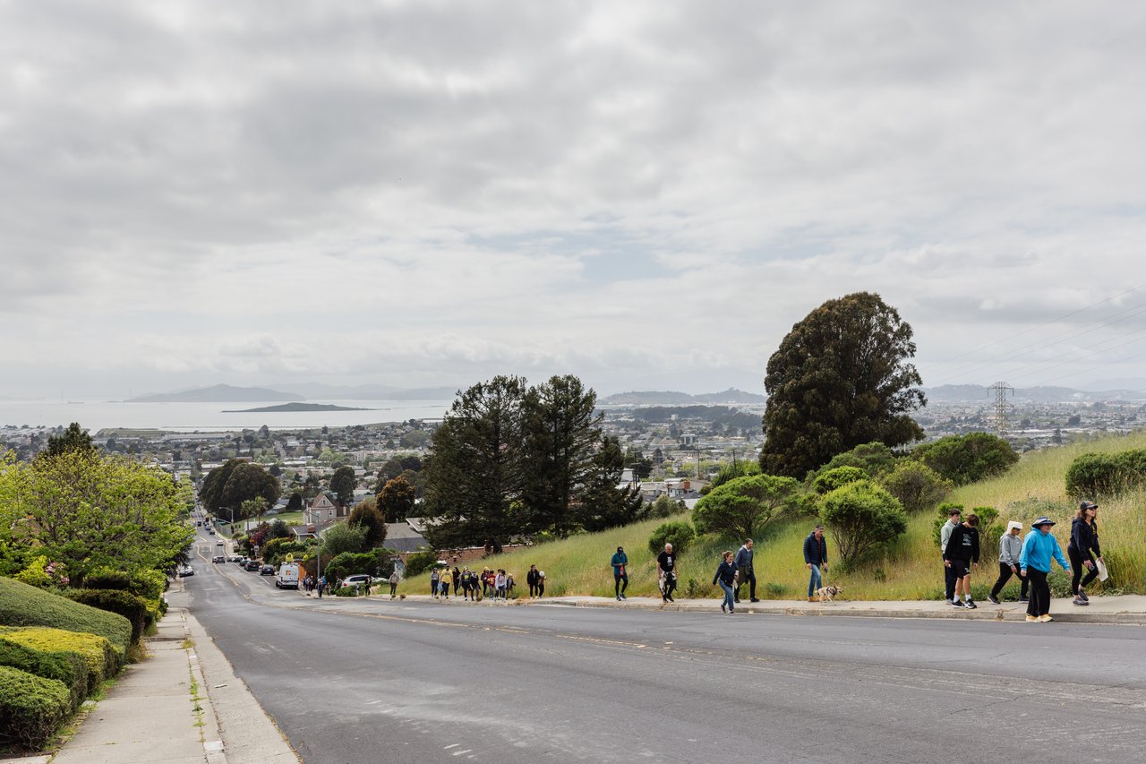 Hillside street overlooking a residential neighborhood, the bay, and distant hills, with pedestrians walking along the slope.