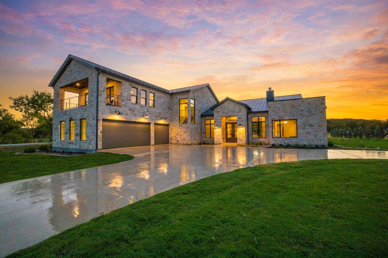 A large house with a garage and a driveway is silhouetted against a colorful sunset. 