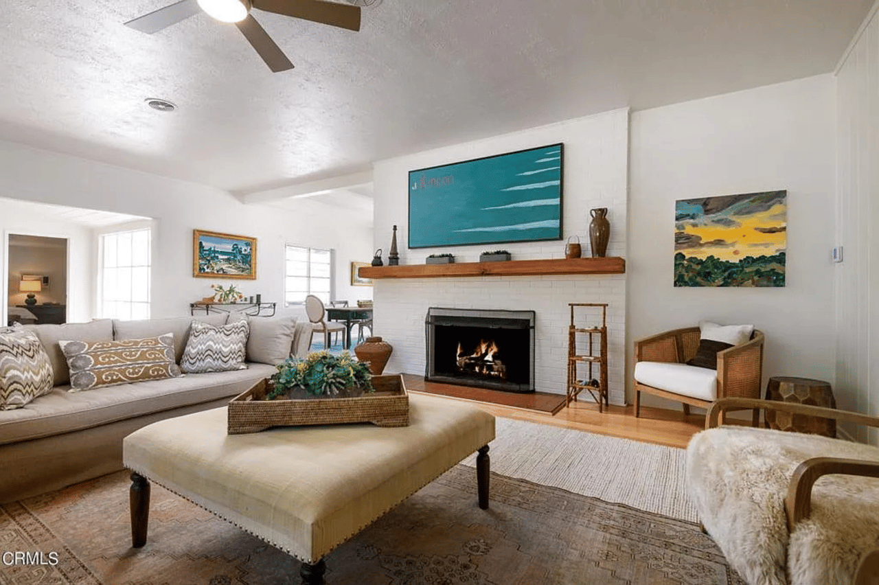 Furnished living room with a white brick fireplace, wooden mantel, and neutral seating.