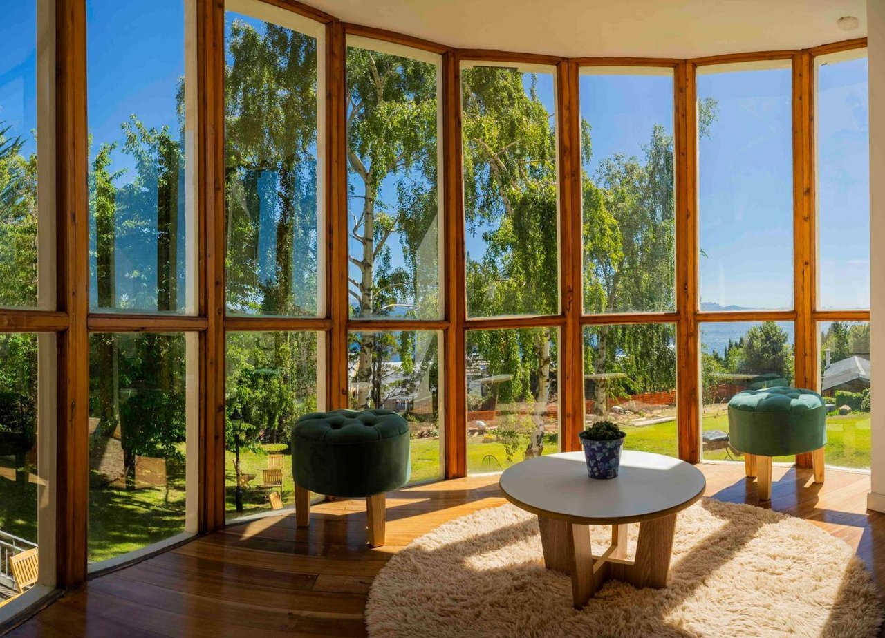Sunroom with floor-to-ceiling windows overlooking a leafy garden, furnished with green ottomans and a round table.