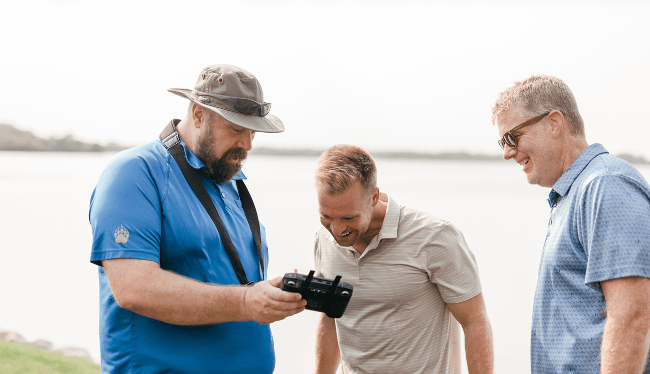 Three men smile as they look at a handheld controller beside the water.