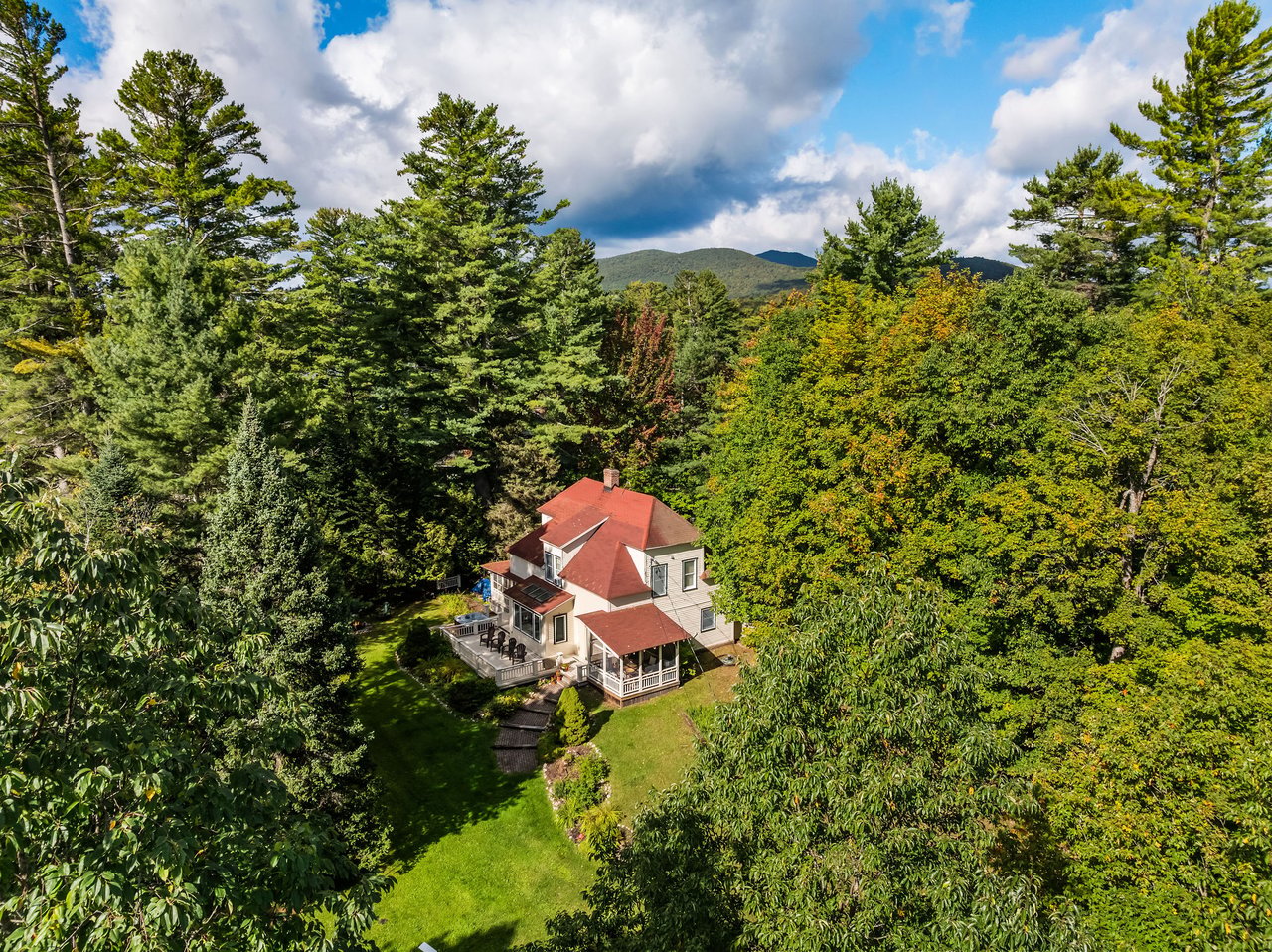 Aerial view of a red-roofed house among dense trees, with a lawn and distant mountains.