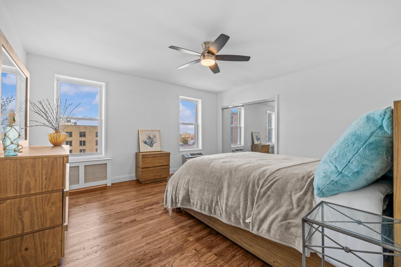 Bedroom with a large bed, wood floors, ceiling fan, and windows overlooking nearby buildings.