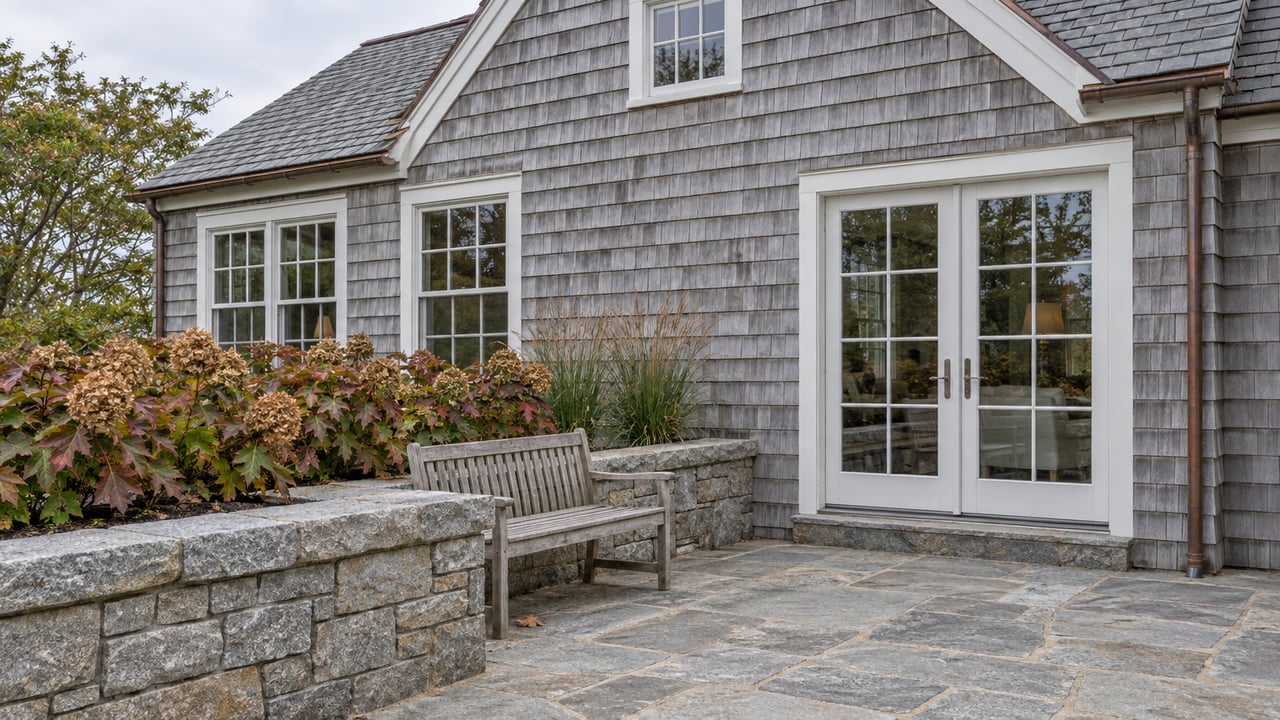 Weathered gray cedar-shingle home with white divided-light windows, a granite terrace, teak bench, and autumn hydrangeas.