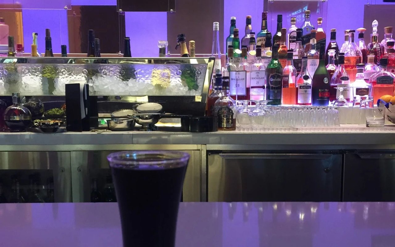 Dark drink on a bar in front of a purple-lit display of liquor bottles.