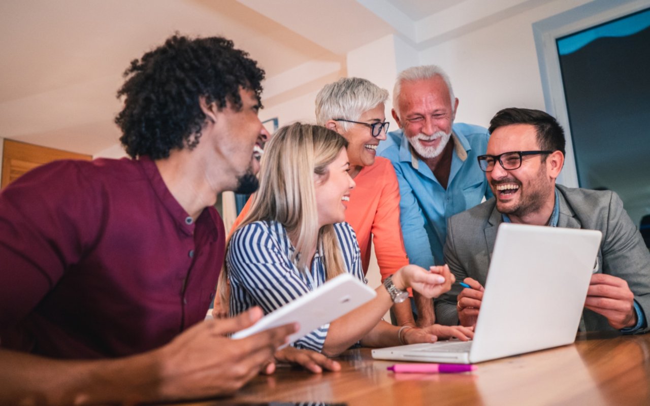 Adults from different generations laugh together around a laptop and tablet at home.