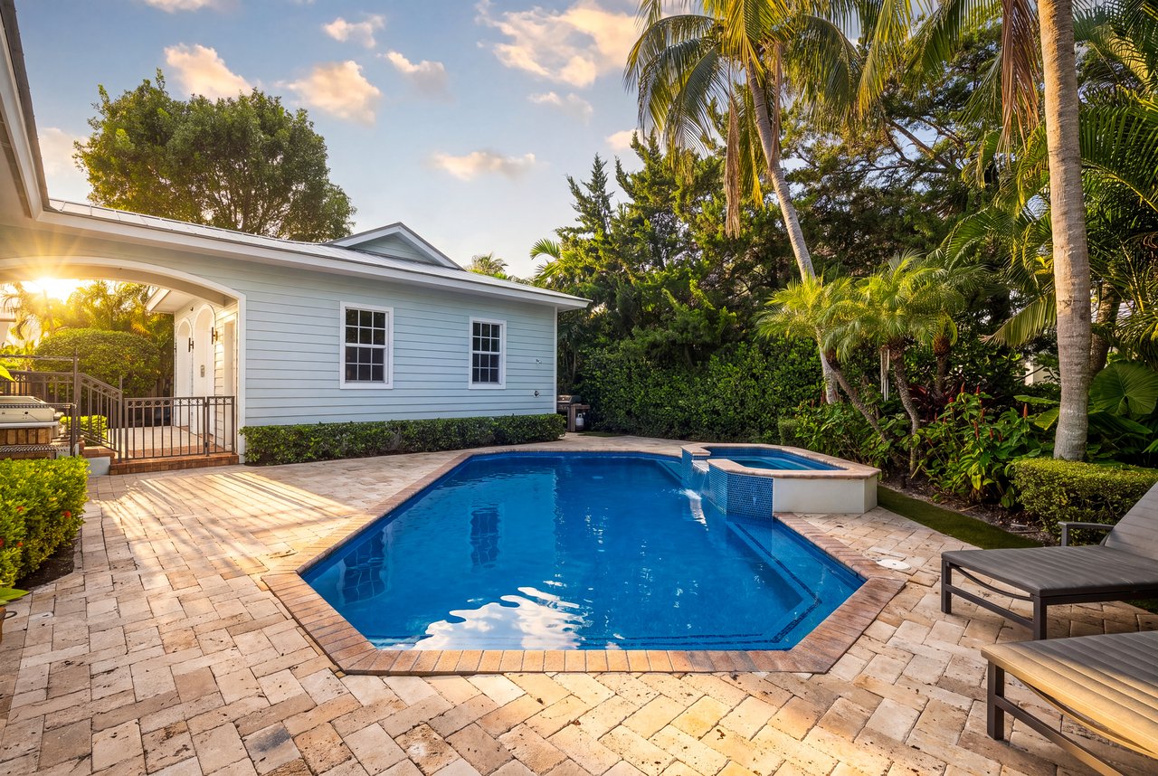 Backyard pool and spa beside a pale blue home, framed by palms at golden hour.