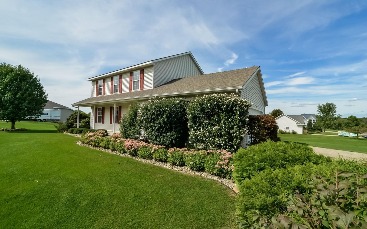 Two-story home with red shutters, landscaped flower beds, and a broad green lawn.
