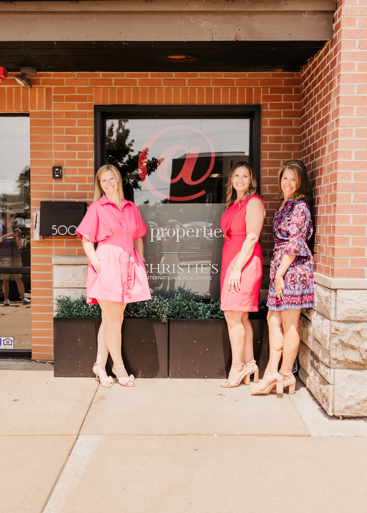 Three women pose outside a brick real estate office, with a large @ symbol on the window.