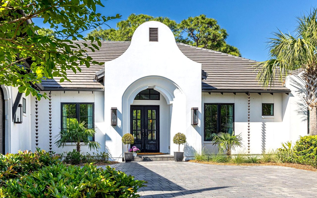 White home with a tall arched entry, dark doors, tropical landscaping, and a paved driveway.