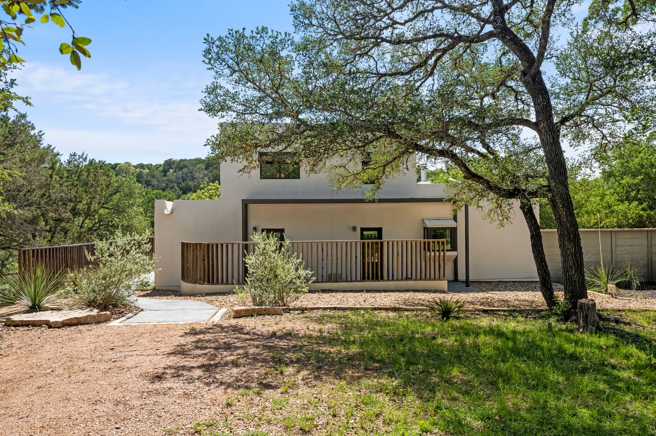 Modern two-story home with a covered patio and wood-slat railing beneath mature trees.