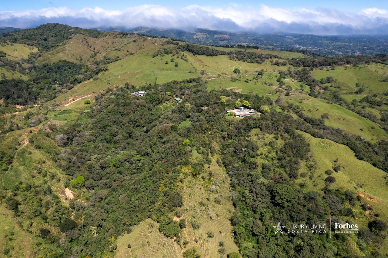 Aerial view of a wooded mountain retreat amid rolling green hills and distant cloud-covered peaks.