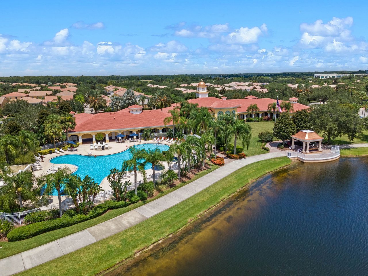 Aerial view of a community clubhouse and swimming pool beside a lake, surrounded by palm trees and homes.