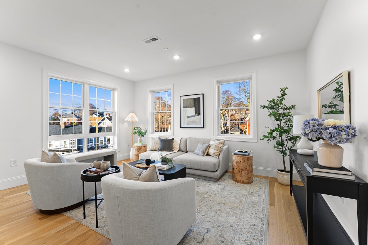 Furnished living room with light sofas, hardwood floors, and tall windows overlooking autumn trees.