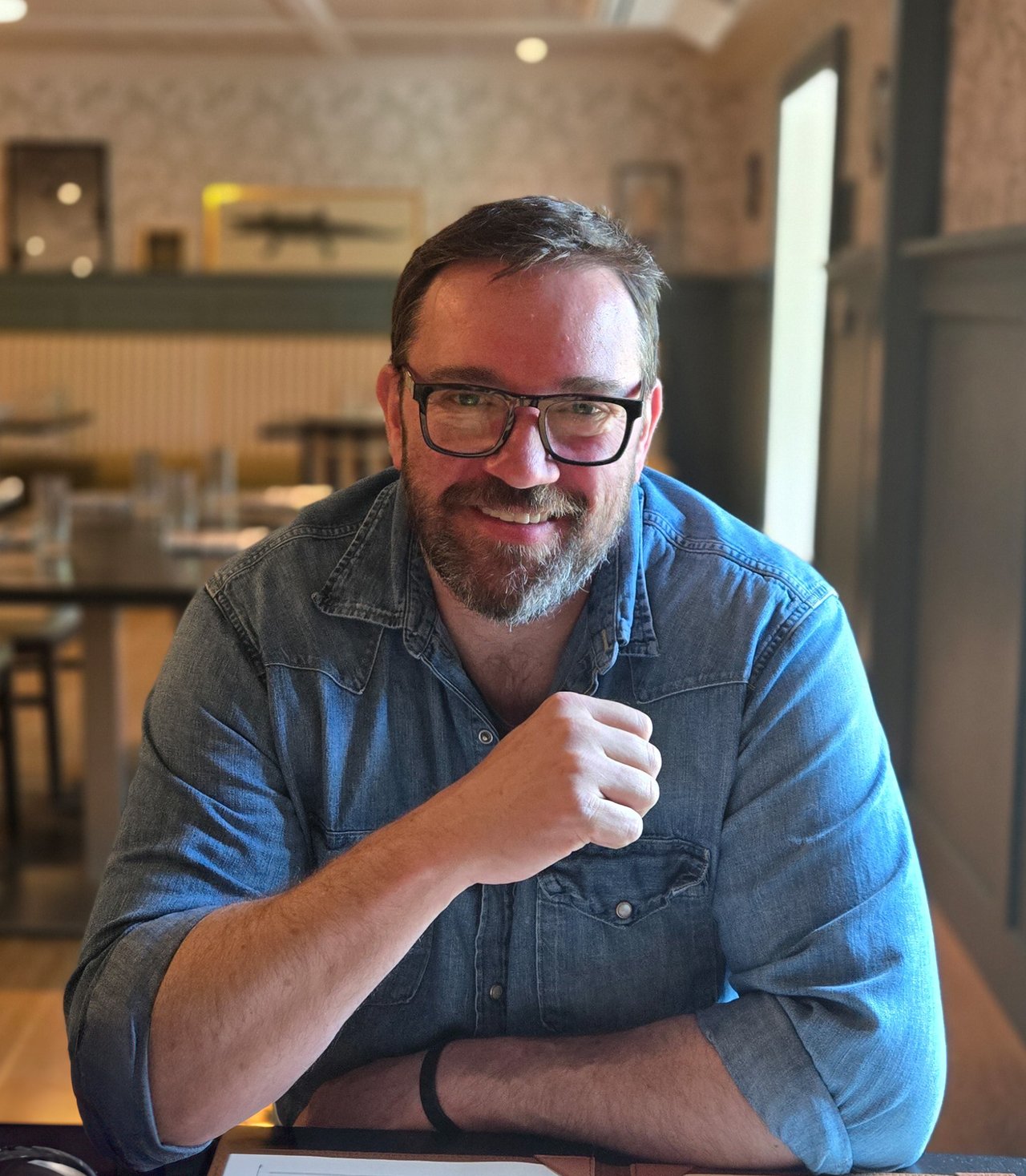 Smiling bearded man in glasses and a blue denim shirt seated at a café table.