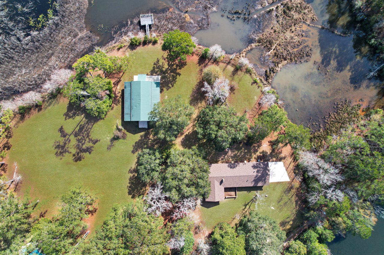 Aerial view of a lakeside property with two buildings, one with a green roof, surrounded by trees and a lawn. A dock extends into the lake, conveying tranquility.