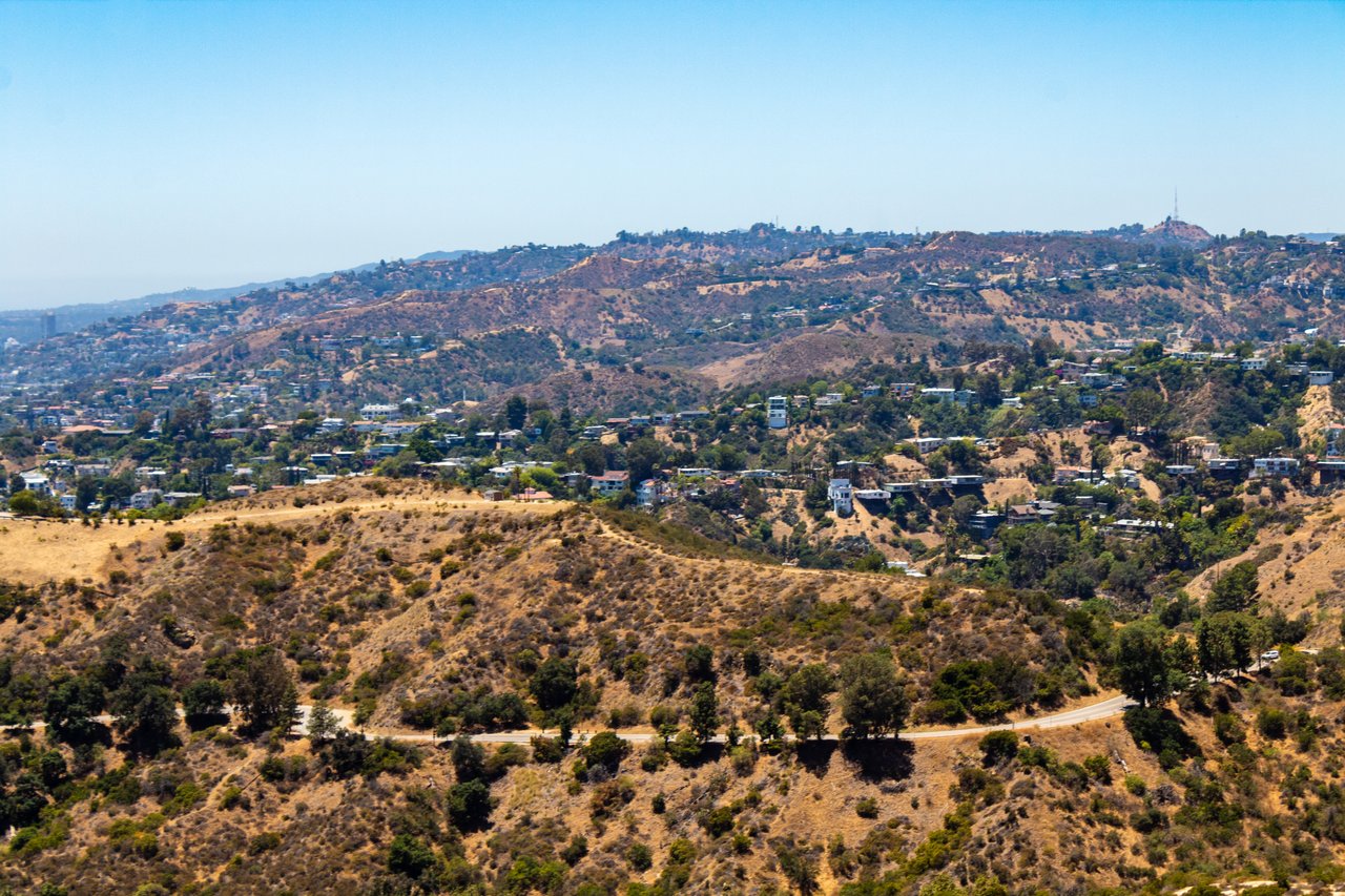 Dry hills dotted with homes and trees, viewed across a winding road beneath a clear sky.