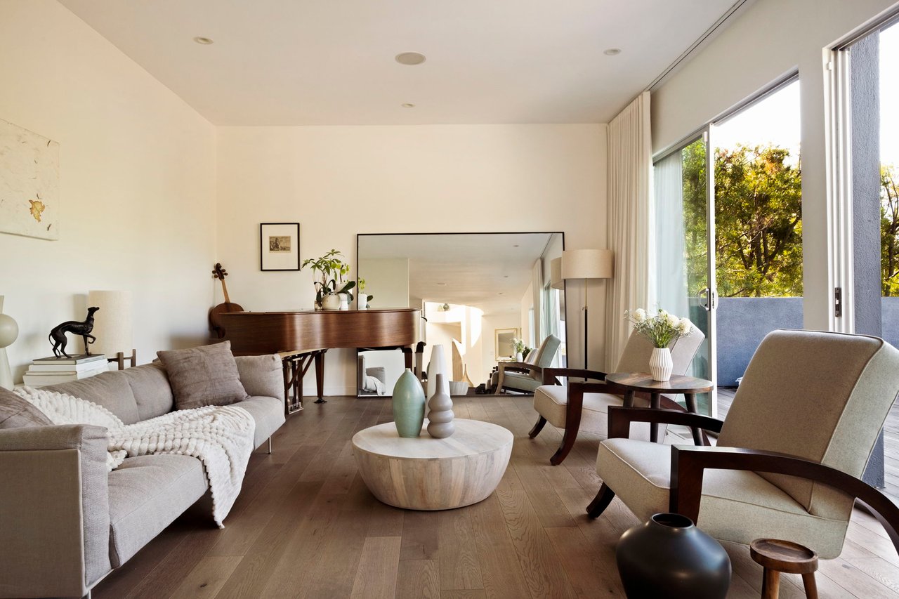 Living room with a grand piano, neutral seating, and large windows overlooking leafy trees.