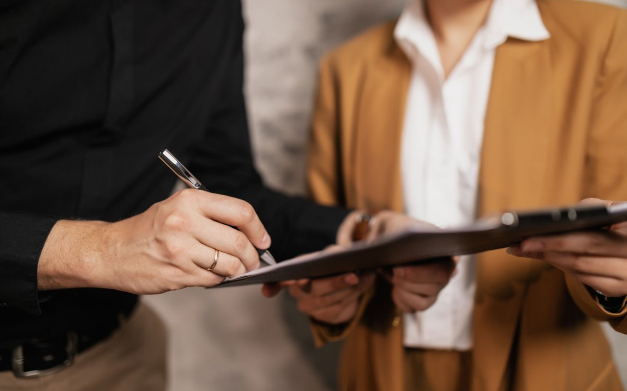 Two people sign a clipboard, one wearing a black jacket and the other a tan blazer.
