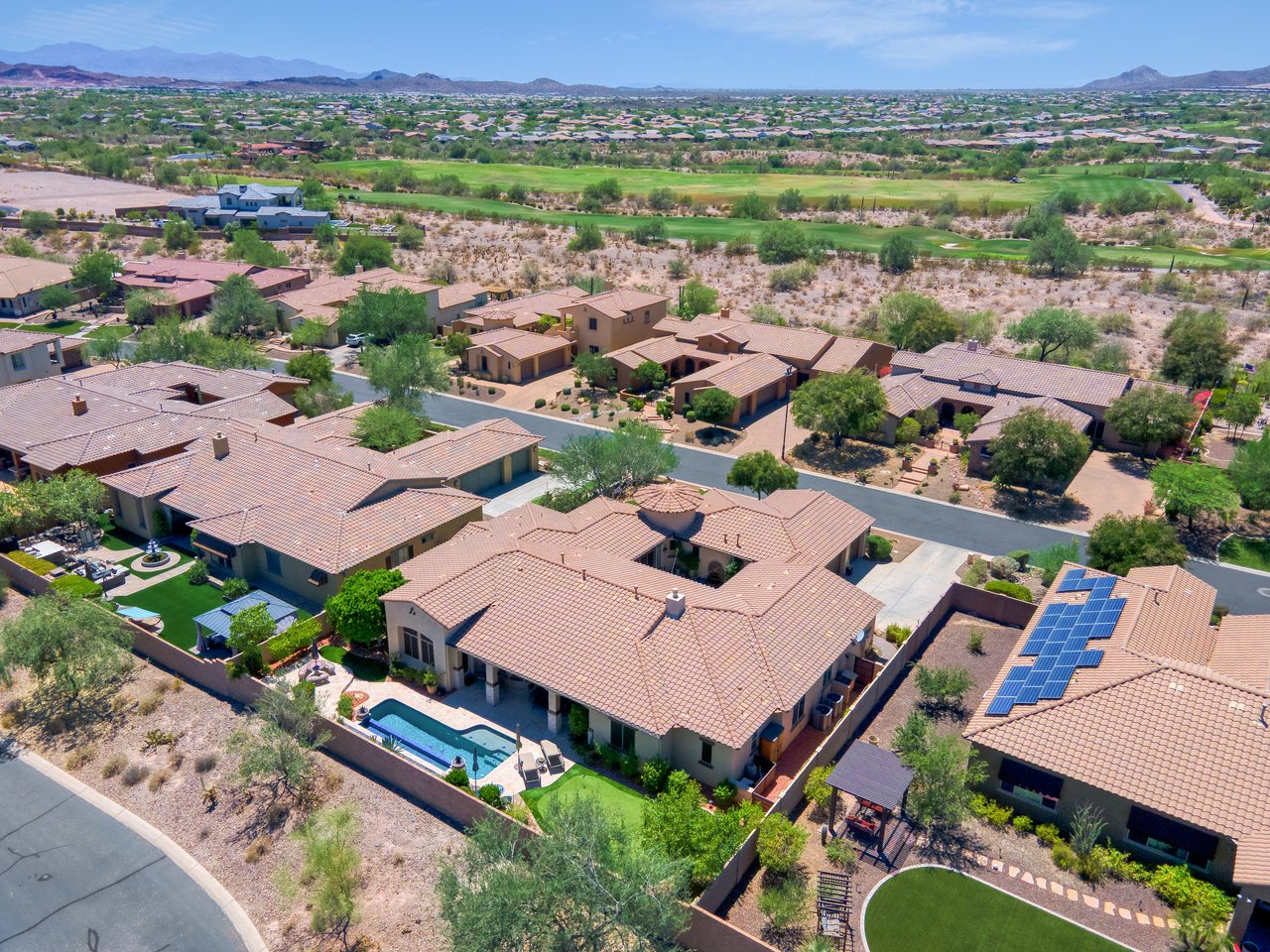 Aerial view of a suburban desert neighborhood with tile-roof homes, a golf course, mountains in the distance, and one backyard pool.