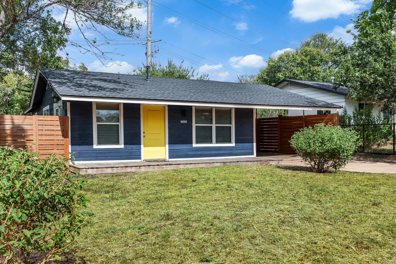 Single-story blue house with a bright yellow front door and fenced front lawn.