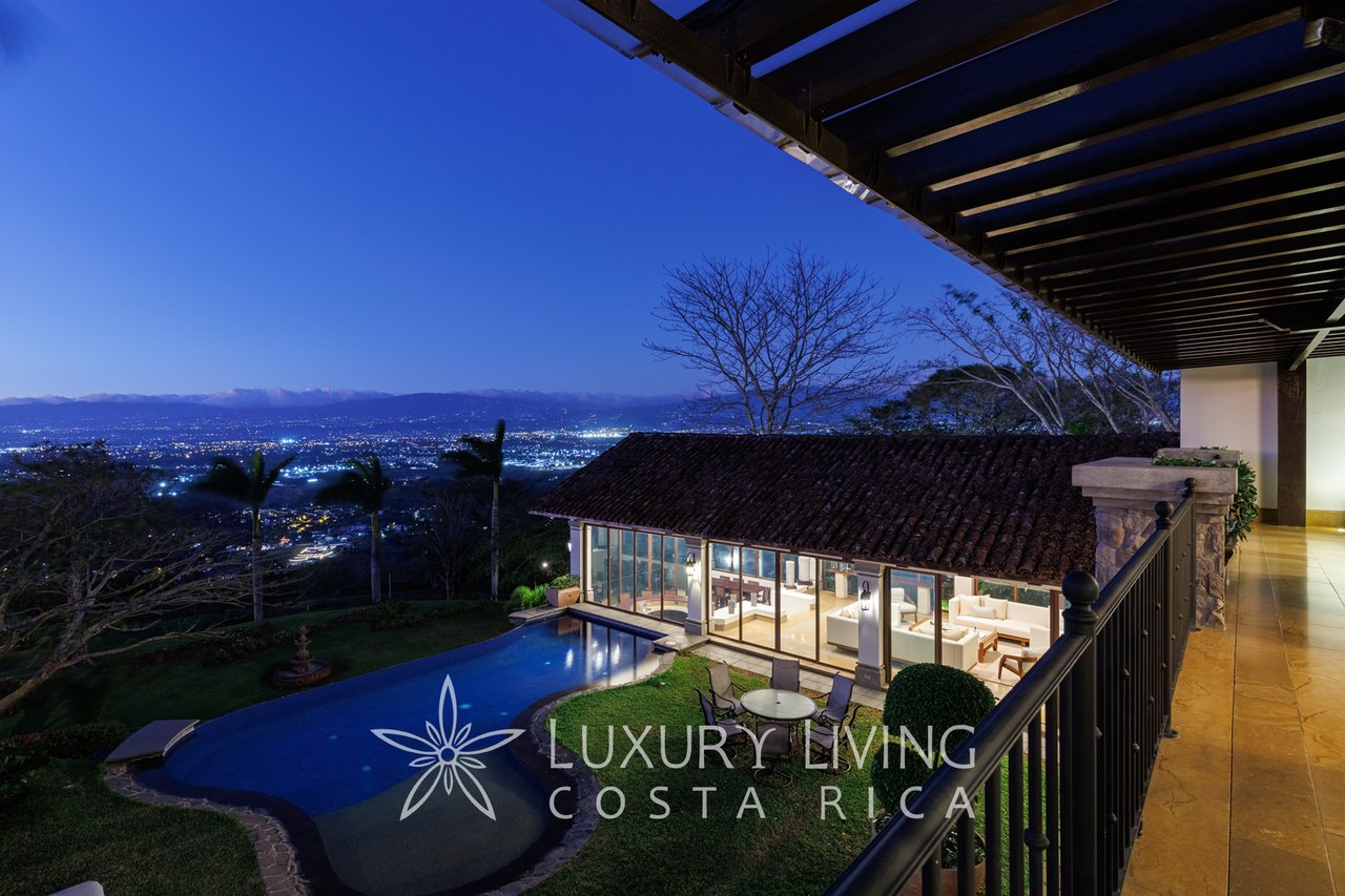 Luxury home and pool overlooking city lights and distant mountains from a covered terrace at dusk.