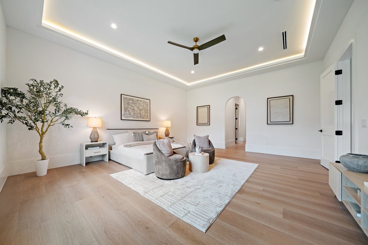 Bedroom with a bed, lounge chairs, light wood floors, and an illuminated tray ceiling.
