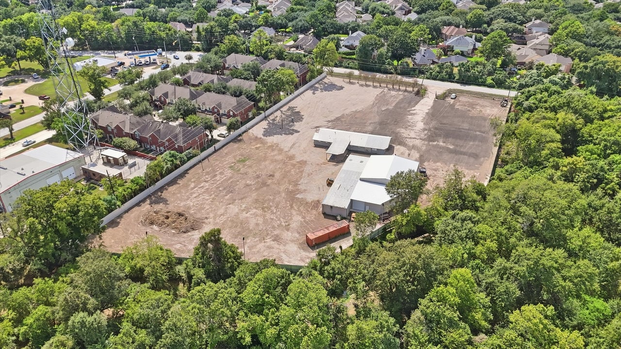 Aerial view of a cleared property with several white-roofed buildings amid trees and nearby homes.