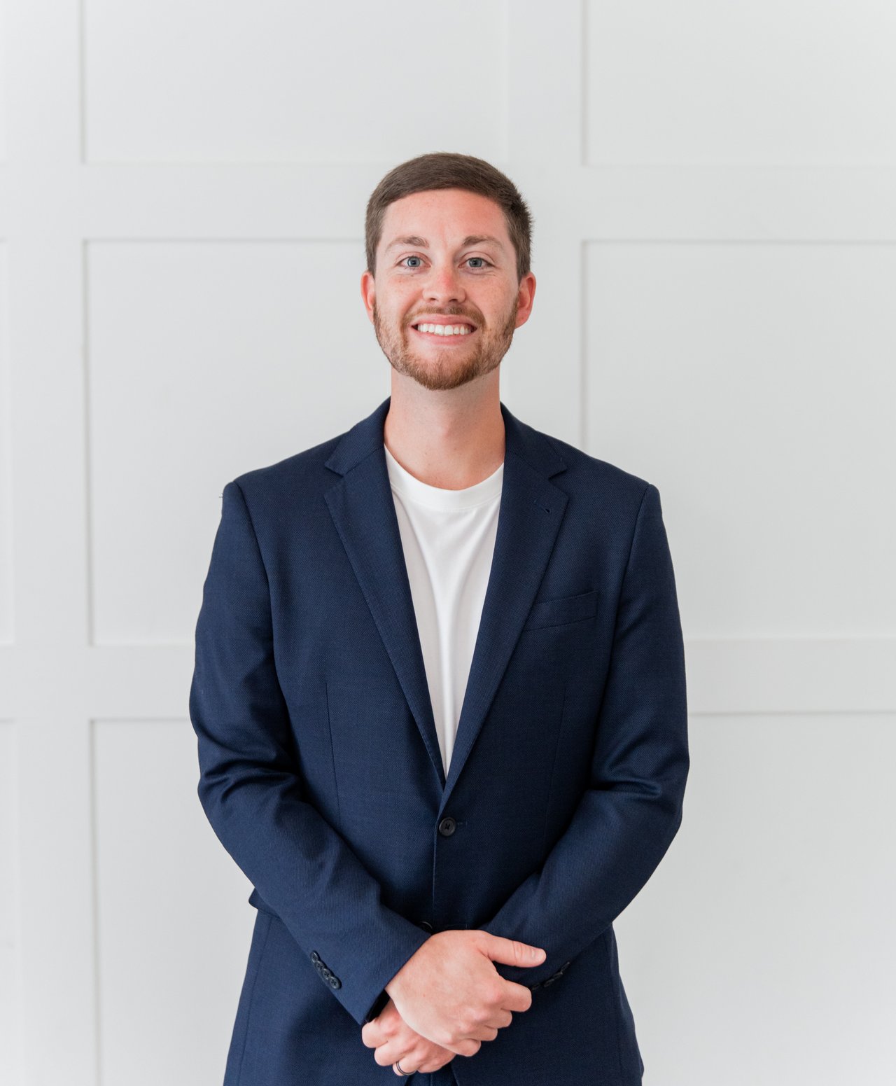 Tommy Barron smiles in a navy suit jacket with his hands clasped against a white paneled wall.