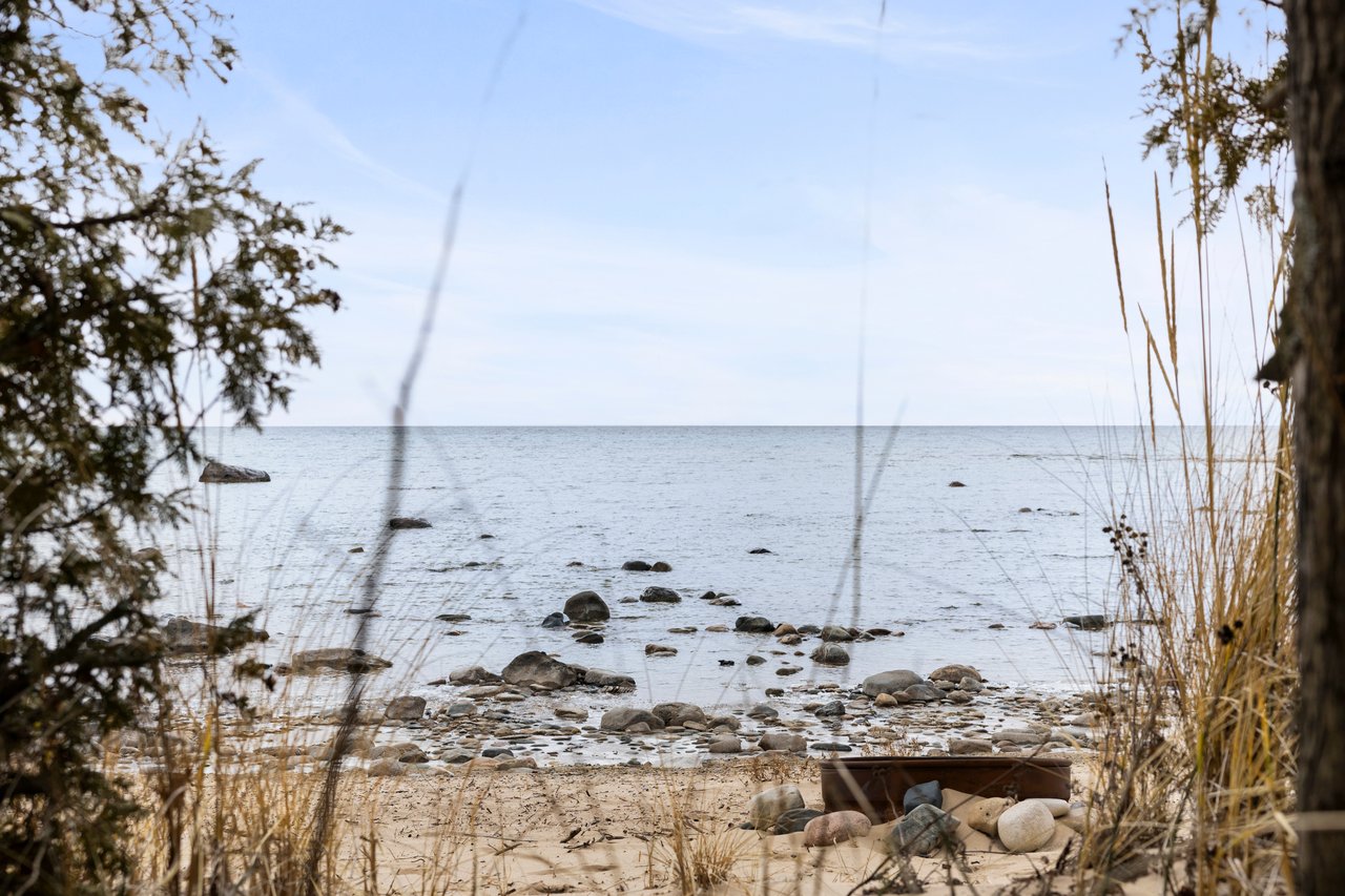 Rocky lakeshore framed by reeds and branches, with calm water beneath a pale blue sky.