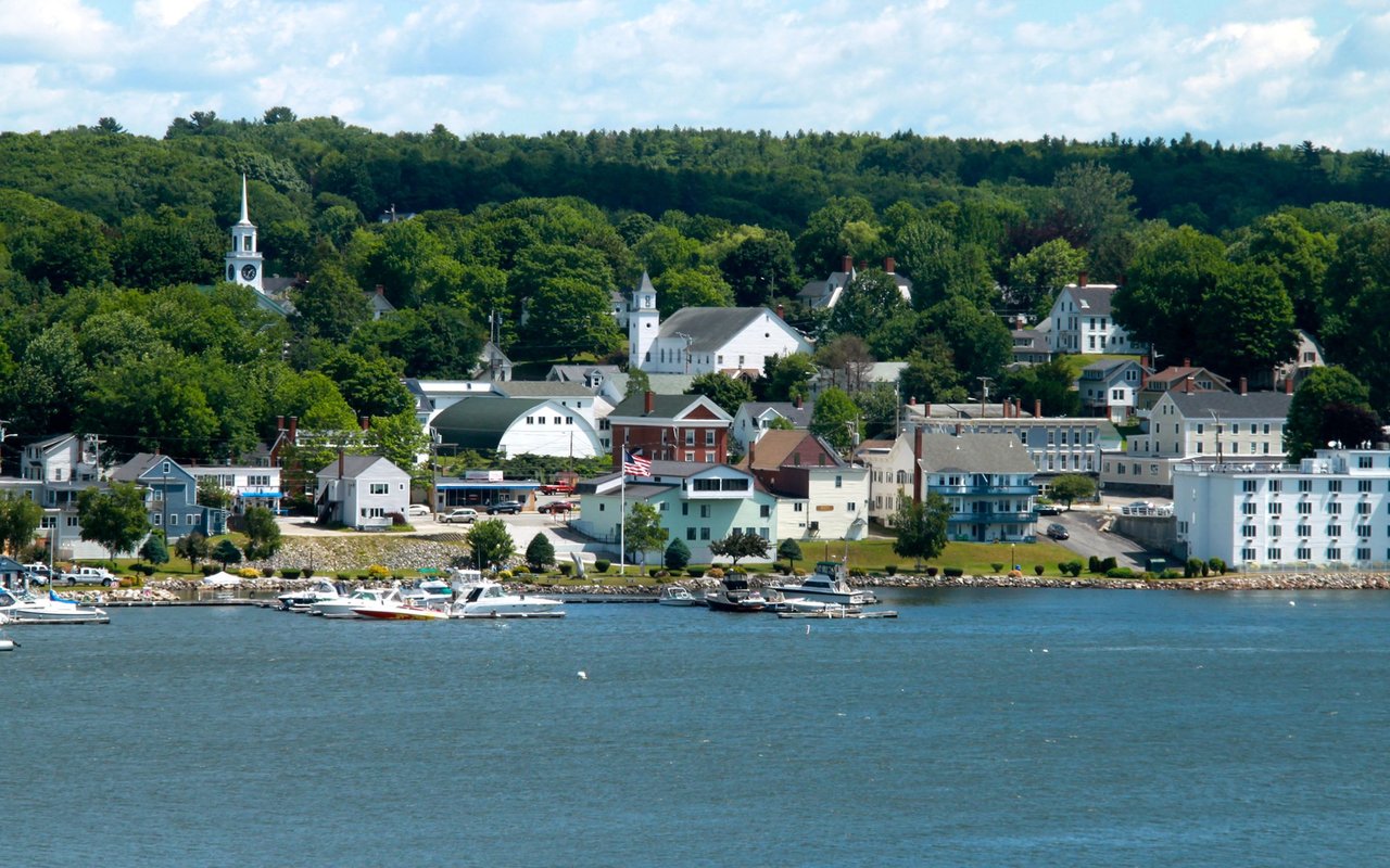 Waterfront town with boats, colorful shorefront buildings, church steeples, and a wooded hill.