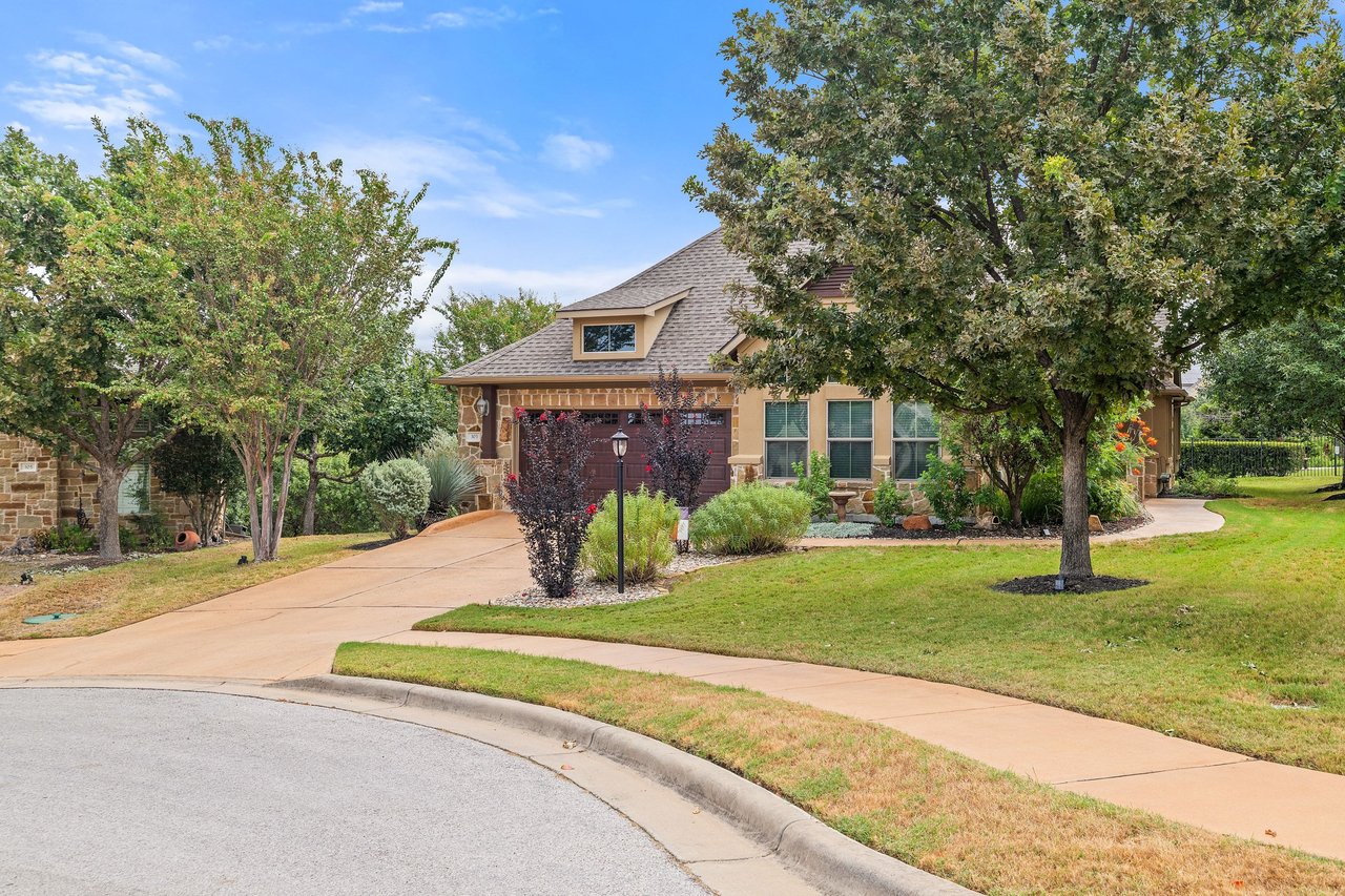 Tan stone-and-stucco home with a curved driveway, landscaped lawn, and mature trees.