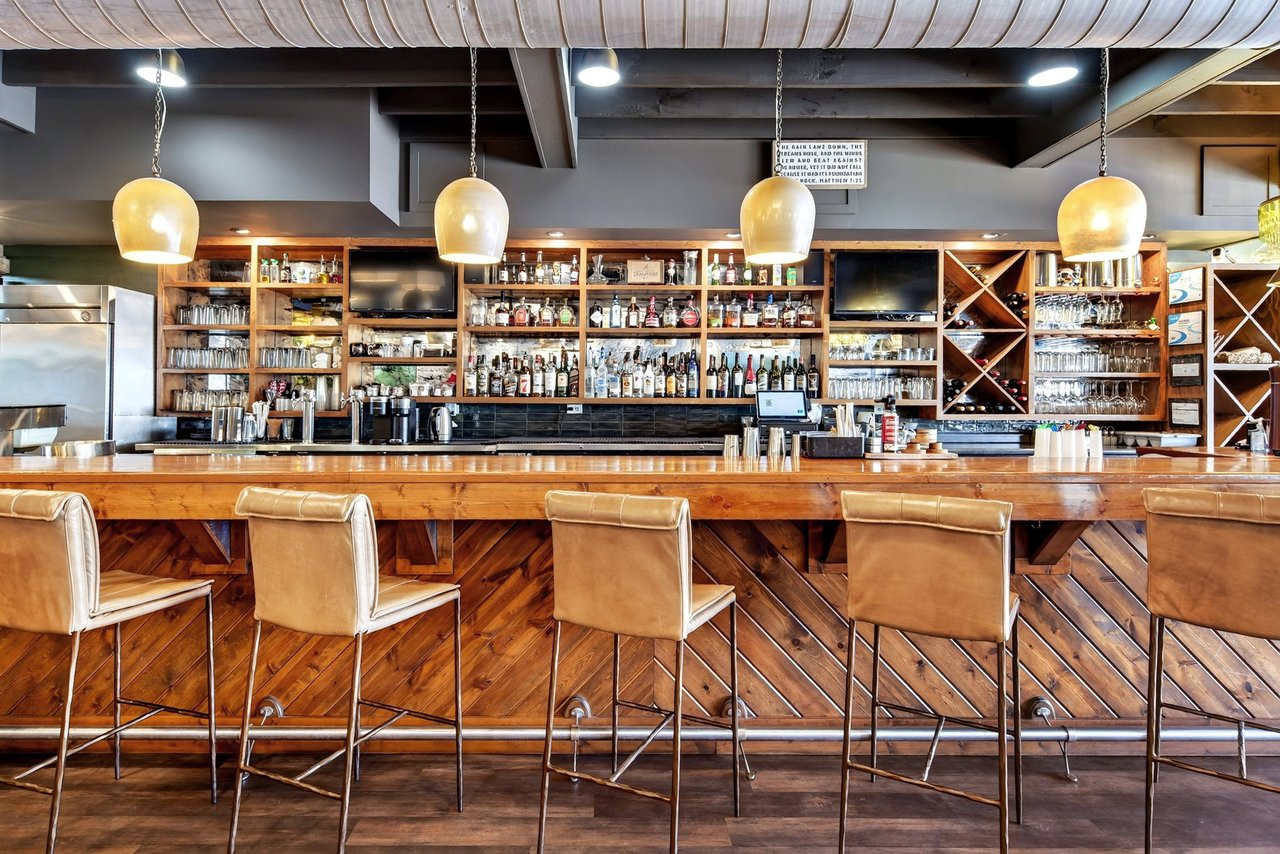 Restaurant bar with a long wooden counter, tan stools, and shelves of bottles and glassware.