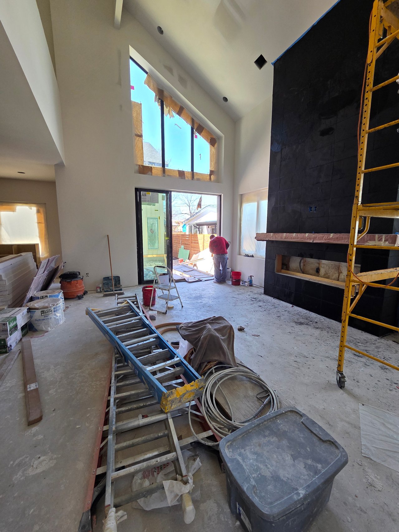 Unfinished living room with tall windows, a dark tiled fireplace wall, and construction equipment.